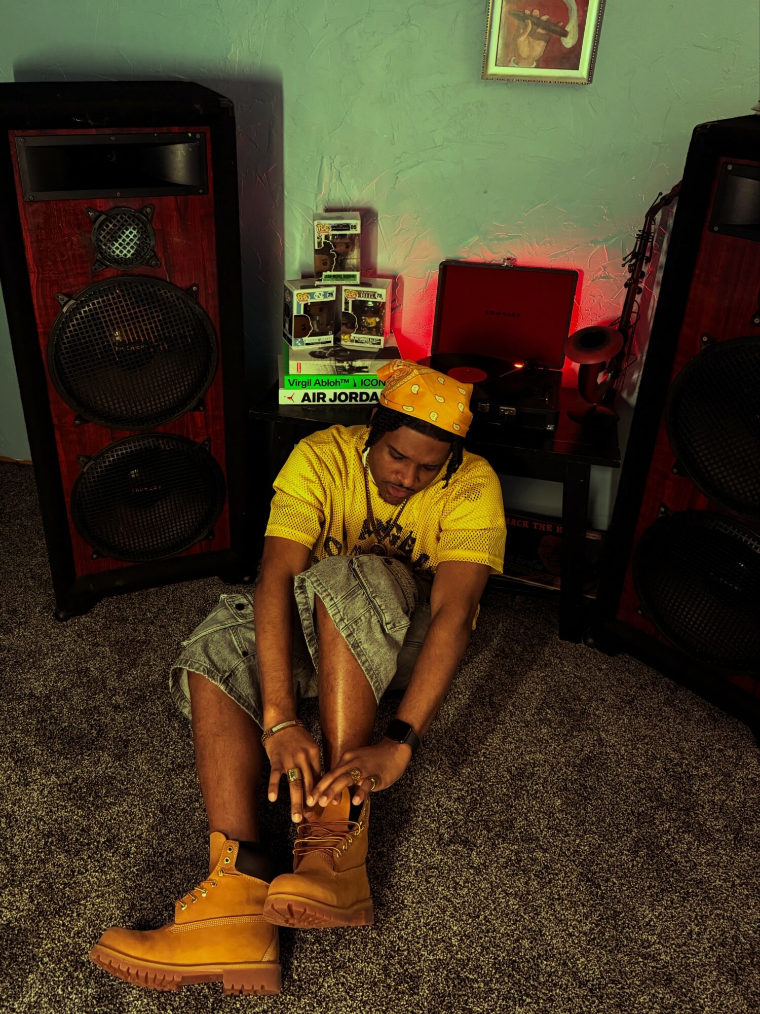Person sitting on the floor wearing yellow shirt, yellow hat, and tan boots, surrounded by speakers and music equipment.