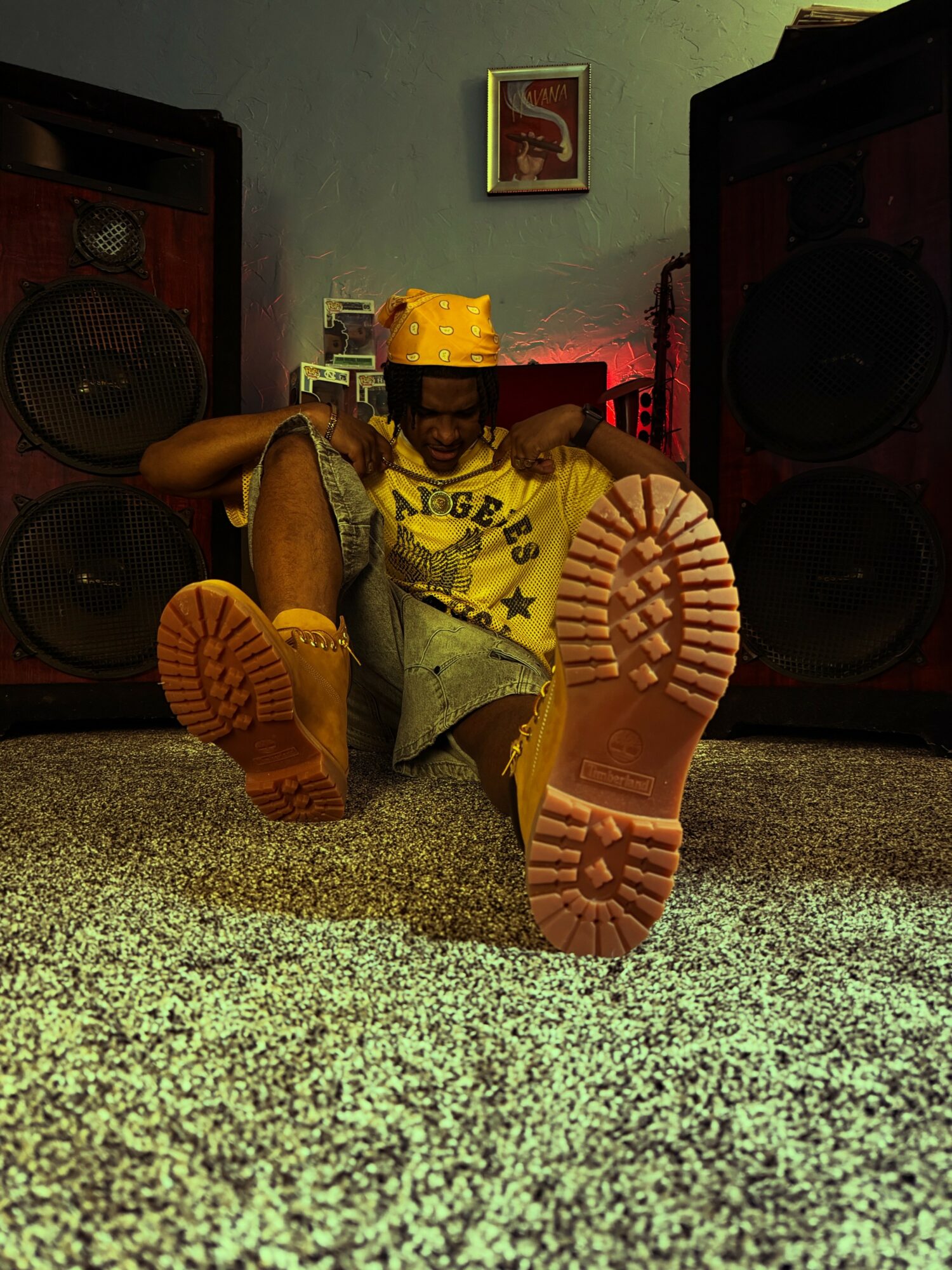 Person sitting on floor with legs extended, wearing yellow shirt and yellow boots, in a room with speakers and framed picture.