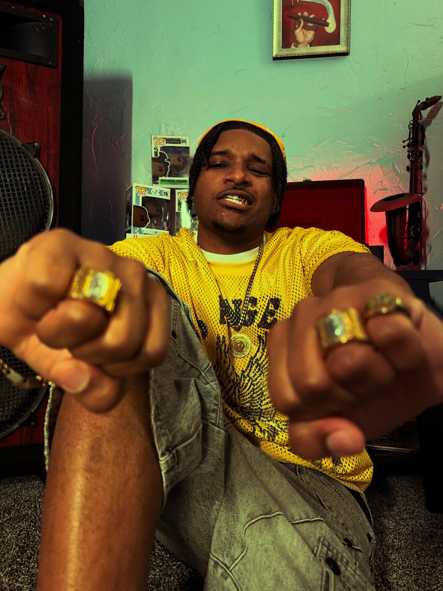 Young man wearing a yellow shirt and jewelry, showing fists towards the camera in a room with green walls.