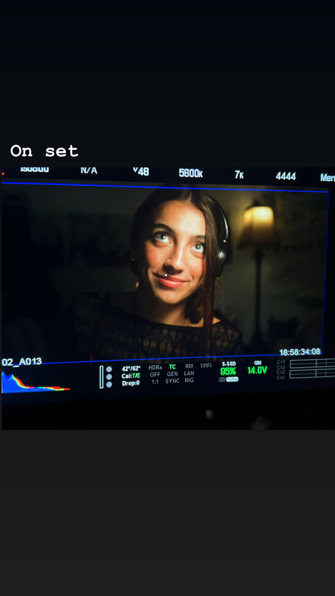 Woman with headphones smiling at camera, on a film or video set, with camera interface overlay visible.