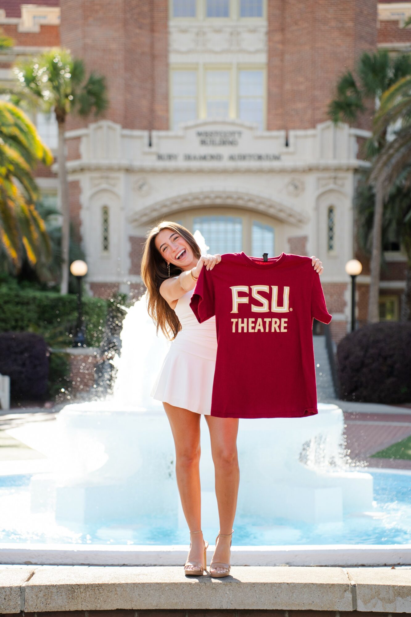 Young woman in white dress holding a red FSU Theatre shirt in front of a fountain and university building.