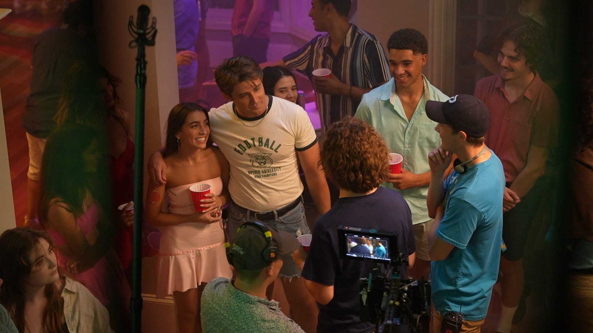 Group of people at a party, some holding red cups, talking and smiling, with a camera recording the scene.