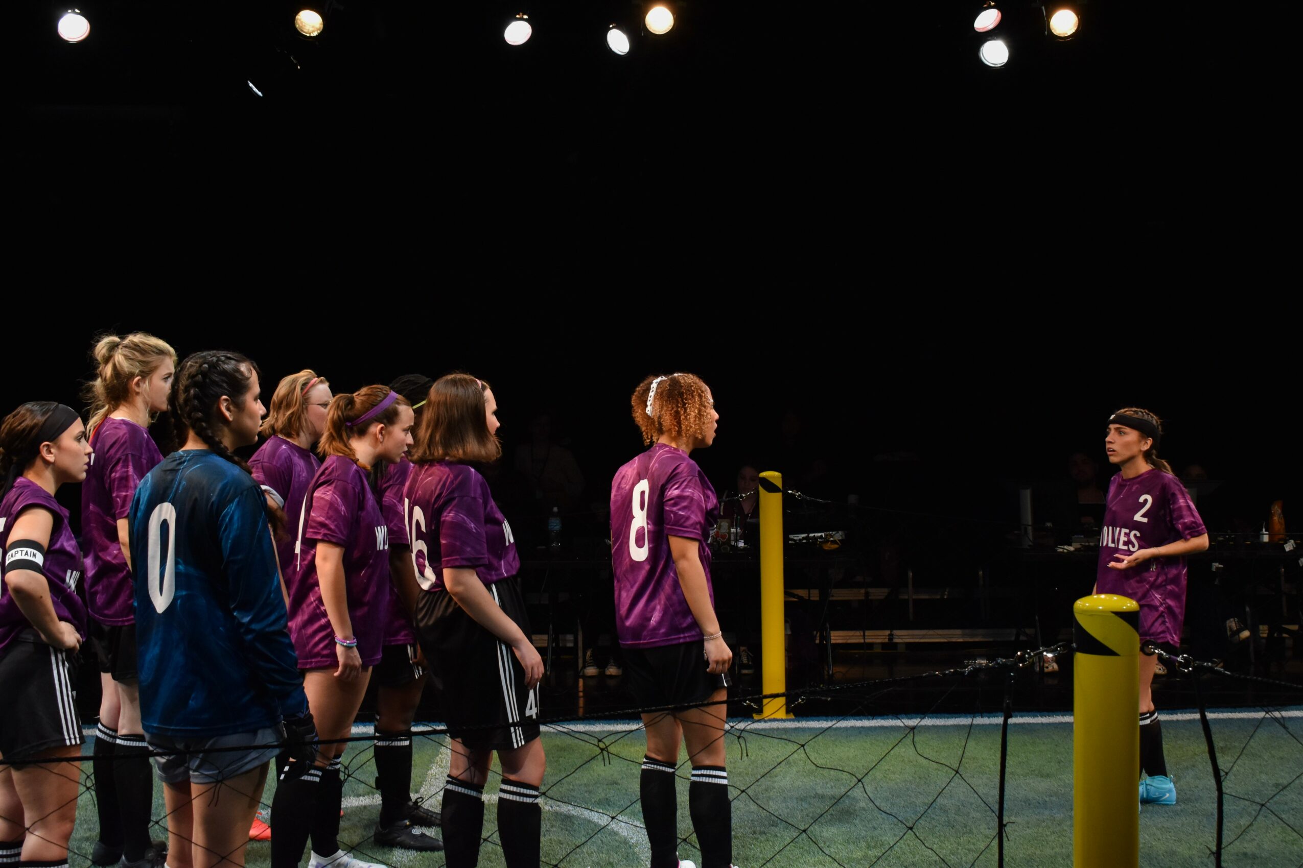 Group of female volleyball players in purple uniforms listening to coach on court, dark background, bright overhead lights.