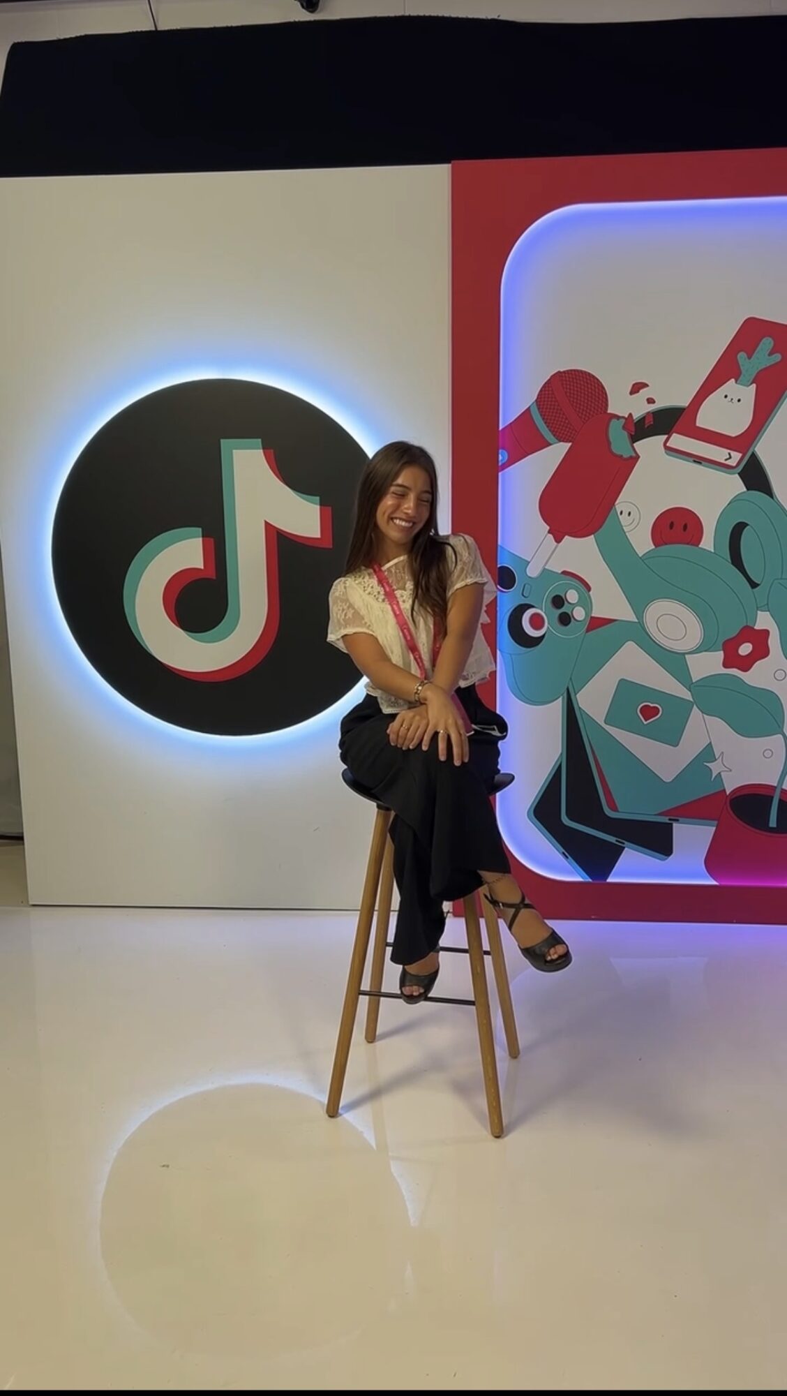 Woman sitting on a stool in front of TikTok logo and colorful digital illustrations, smiling, indoors.