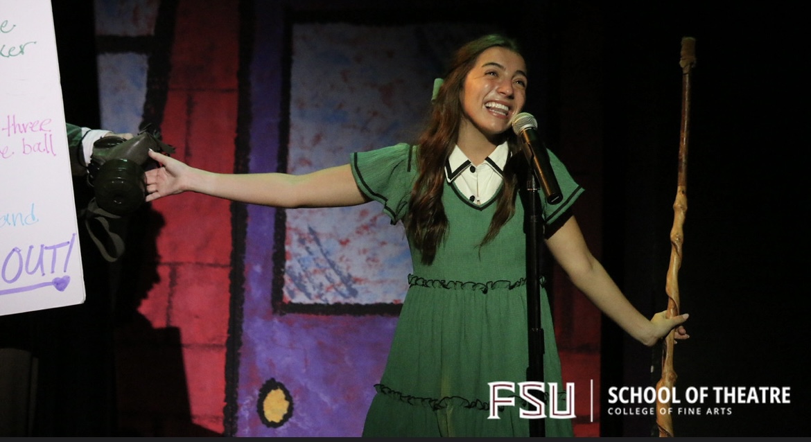 Young woman on stage smiling, holding a microphone and pointing, with a colorful background and a sign, school of theatre.