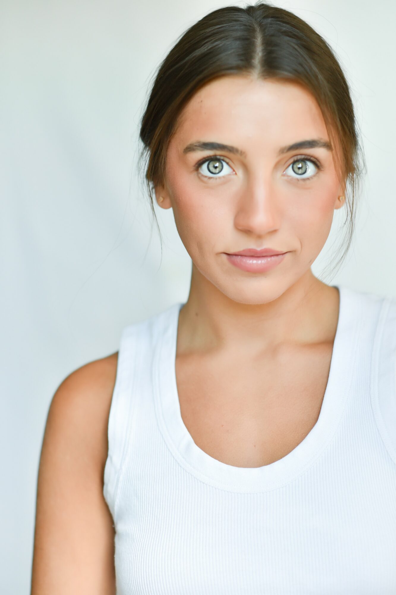 Young woman with dark hair and blue eyes wearing a white sleeveless top, looking directly at the camera.