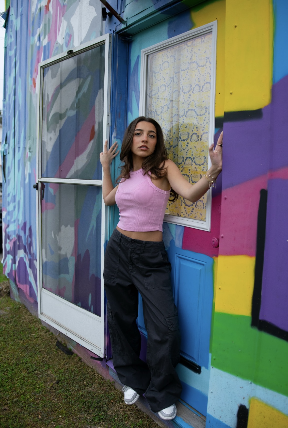 Young woman with long hair in a pink top and dark pants stands in front of a colorful graffiti wall, holding a window frame.
