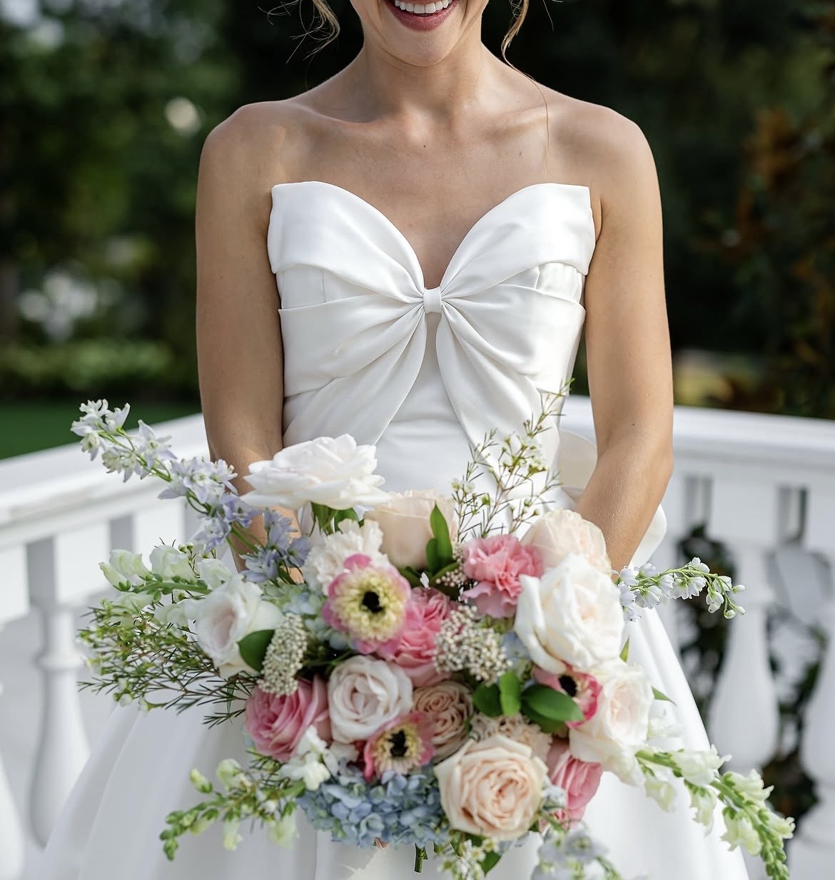 Woman in a strapless white dress holding a large bouquet of pink, white, and blue flowers outdoors.