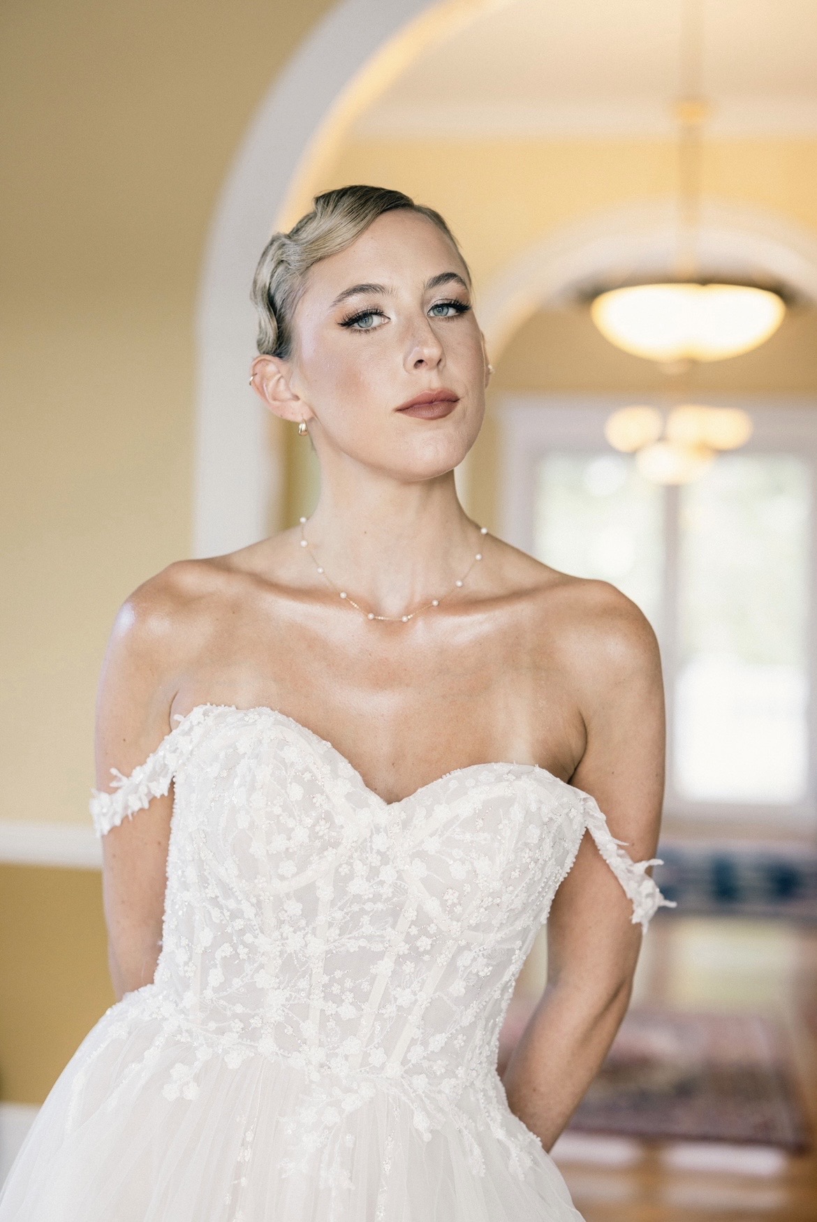 Woman in wedding dress standing indoors with a neutral expression, arched doorway behind her, warm lighting.