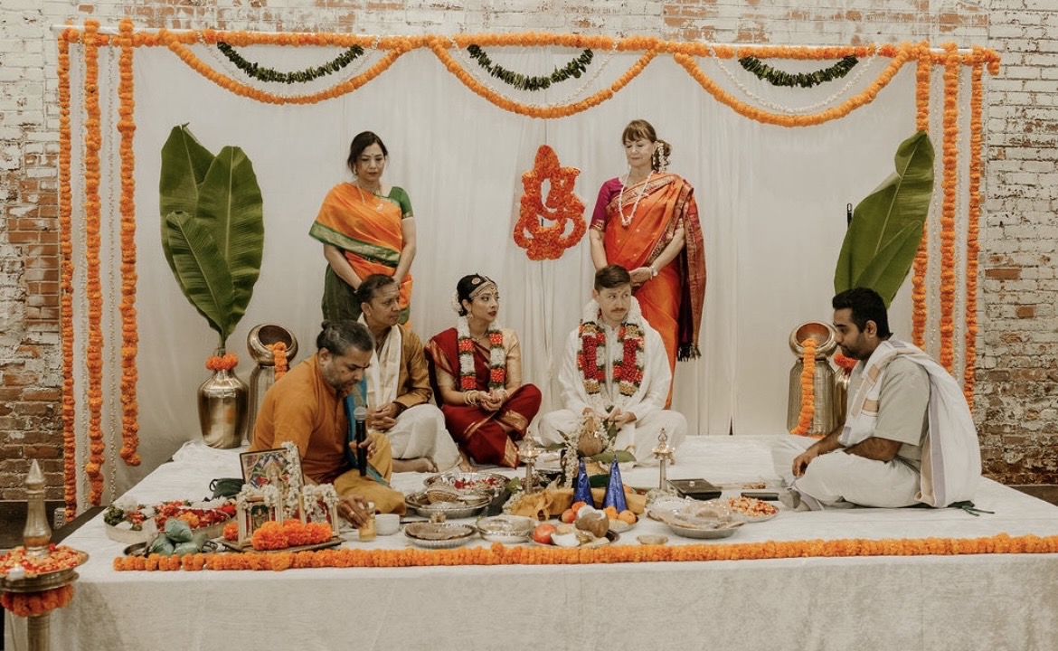 People participating in a traditional ceremony, seated on a decorated platform with floral and fabric decorations, and large green leaves in the background.