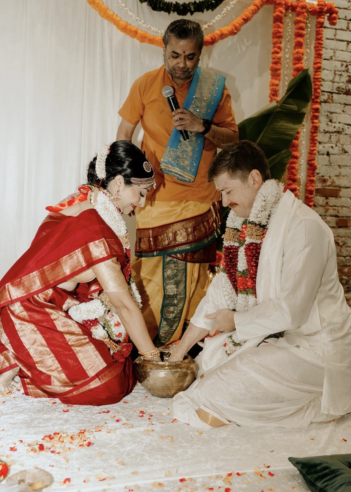 Two people participate in a traditional ceremony, with a man holding a microphone and a woman in a red saree, both kneeling on the floor, with a priest or officiant overseeing the ritual, decorated with orange marigold flowers and green leaves.