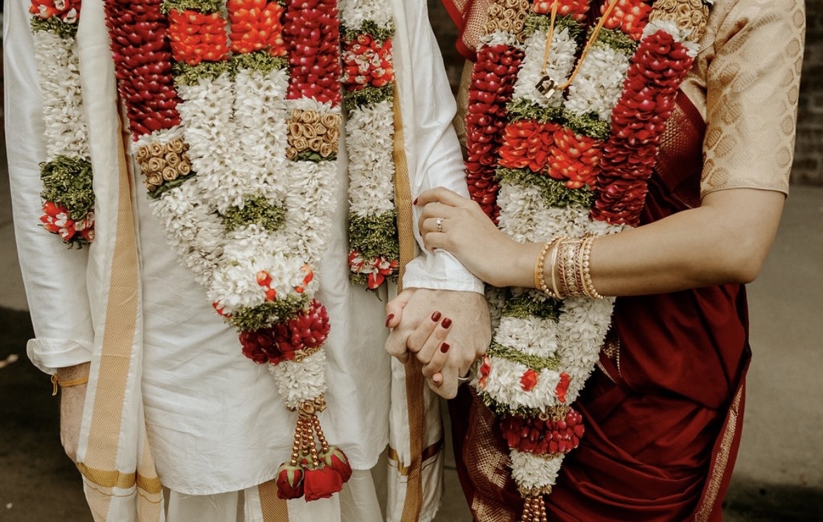 Two people wearing traditional attire with floral garlands, holding hands, and jewelry visible on one person's arm.