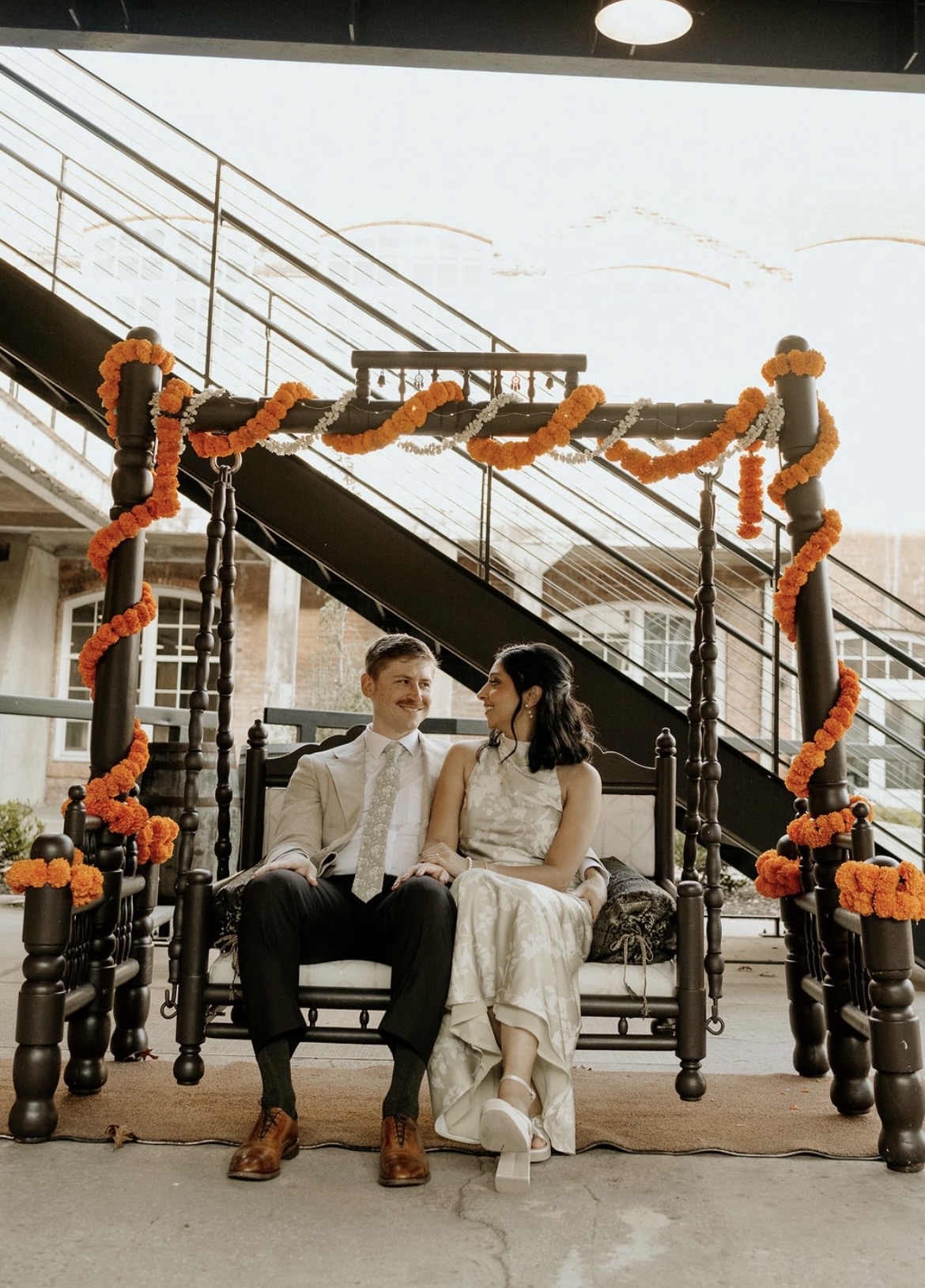 A couple sitting on a decorated swing with orange and black garlands, indoors with a staircase in the background.