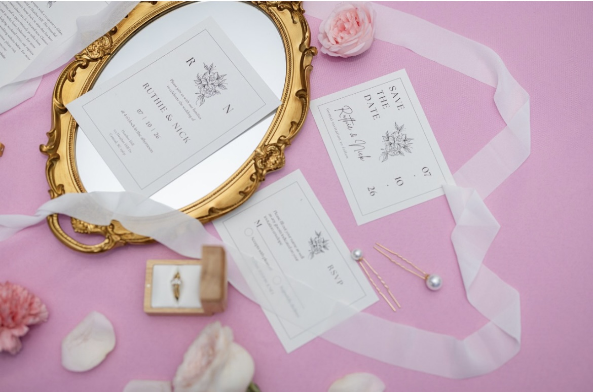 Wedding invitation, pearl earrings, pink flowers, and ribbon on pink background, with a gold mirror frame.