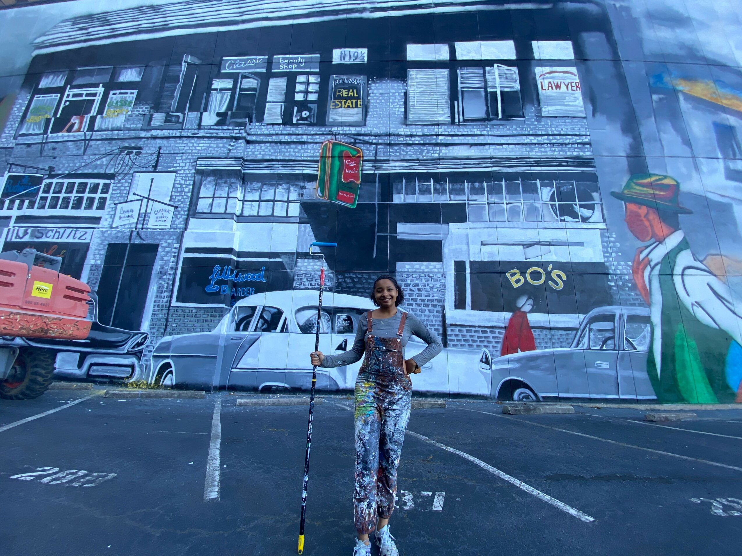Person standing in front of a large mural of a city street scene with buildings, cars, and a person in a hat.