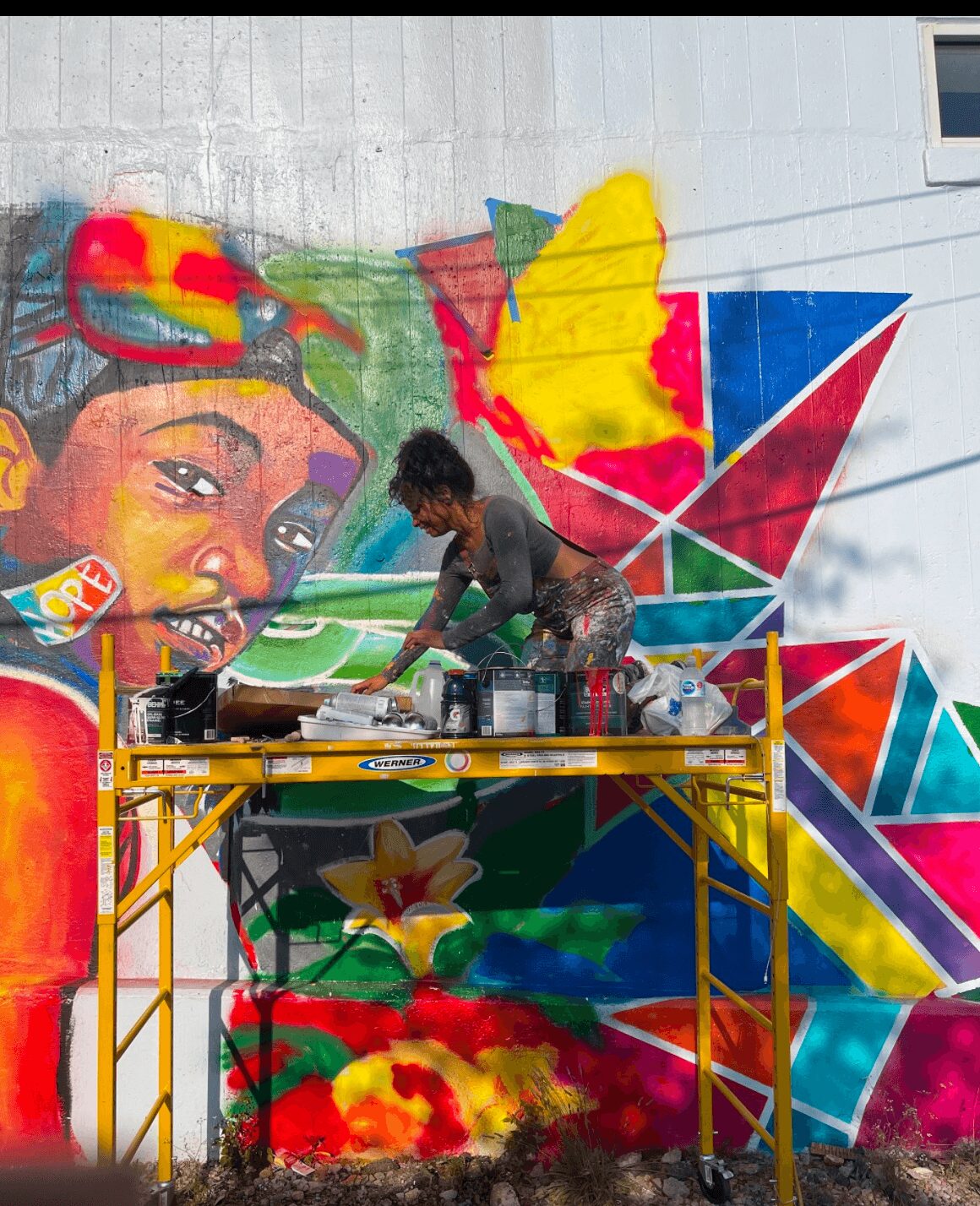 Person working on colorful mural with a large face, geometric shapes, and vibrant flowers, on a wall.