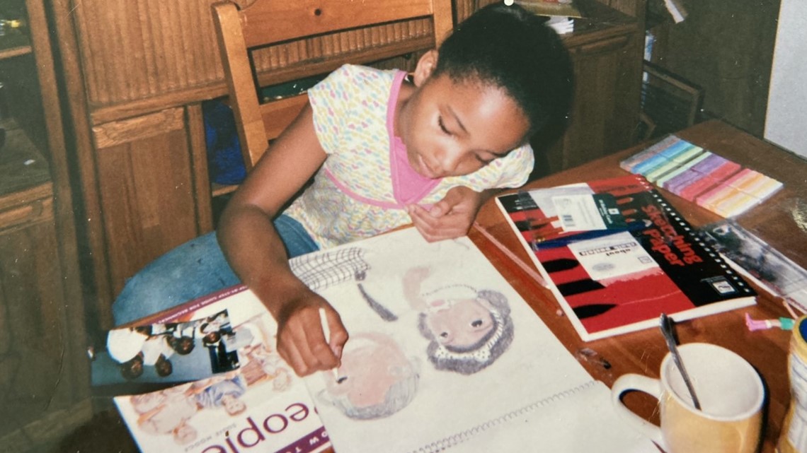 Girl with dark hair in a ponytail, wearing a light-colored shirt, drawing at a table with art supplies and a cup.
