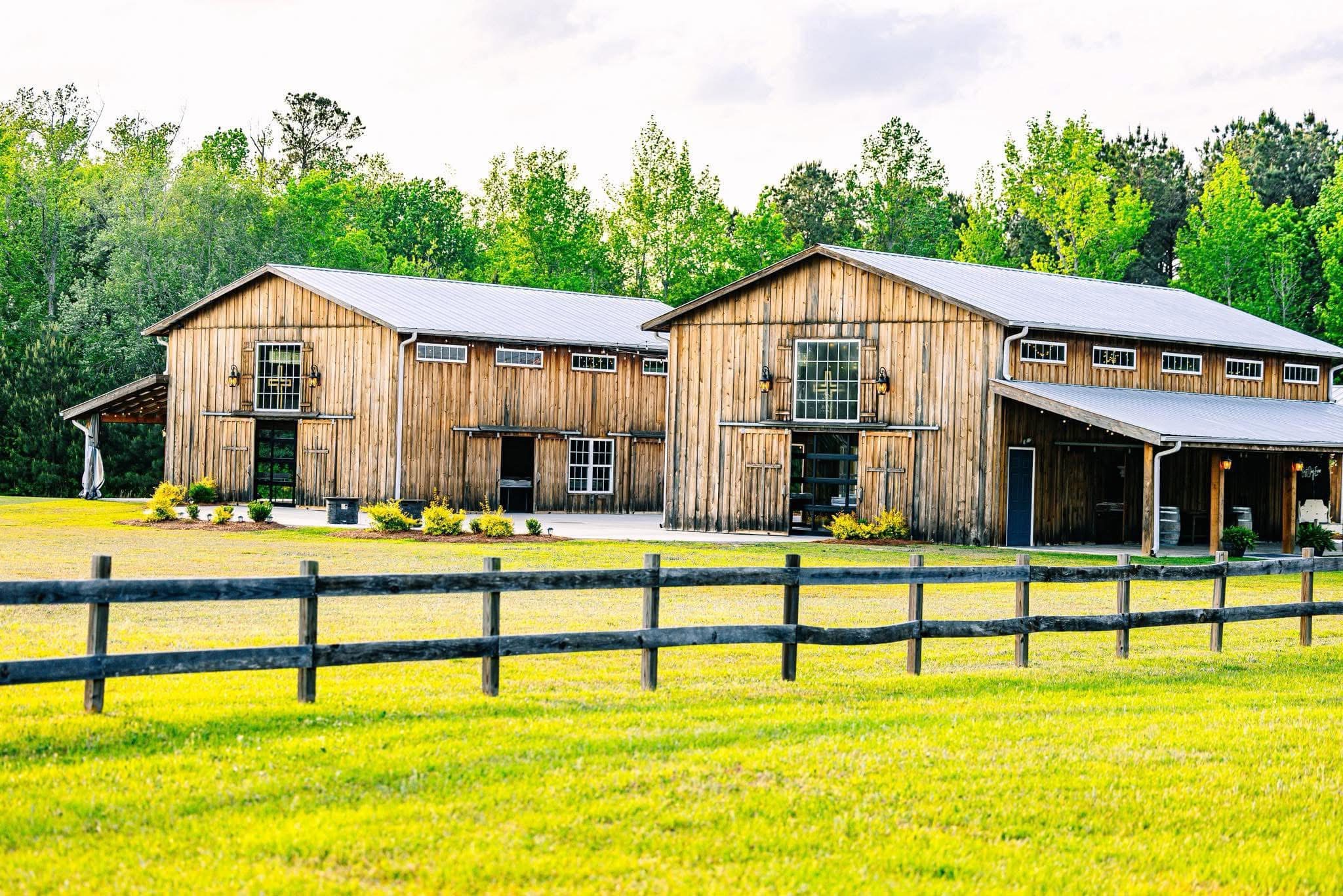 Two wooden barns with metal roofs are in a grassy field, surrounded by a wooden fence and trees in the background.
