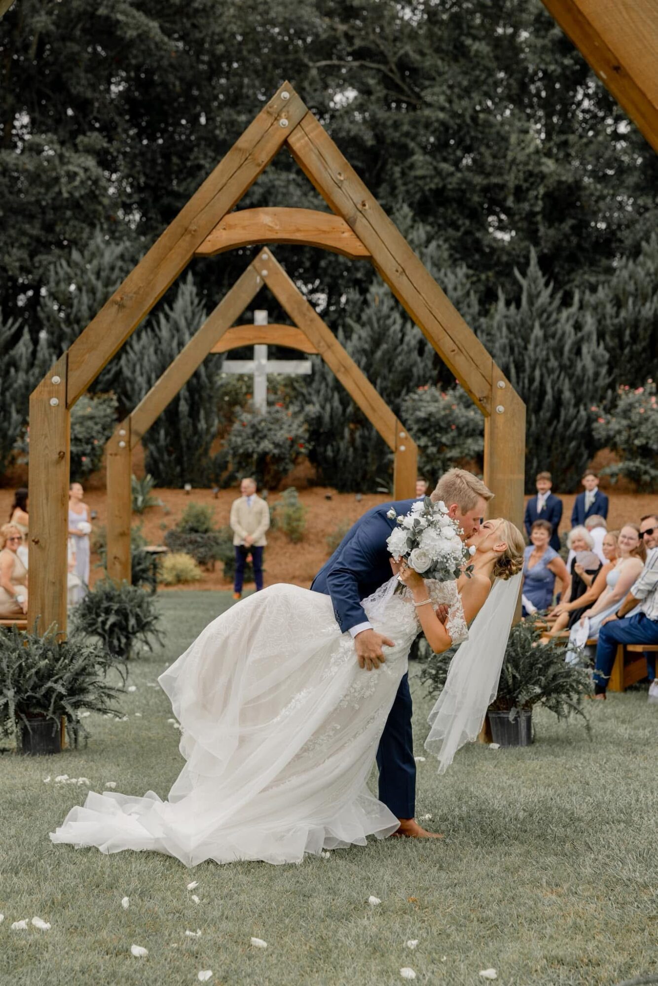 Bride and groom dancing outdoors with guests, wooden arch structures, and trees in background.