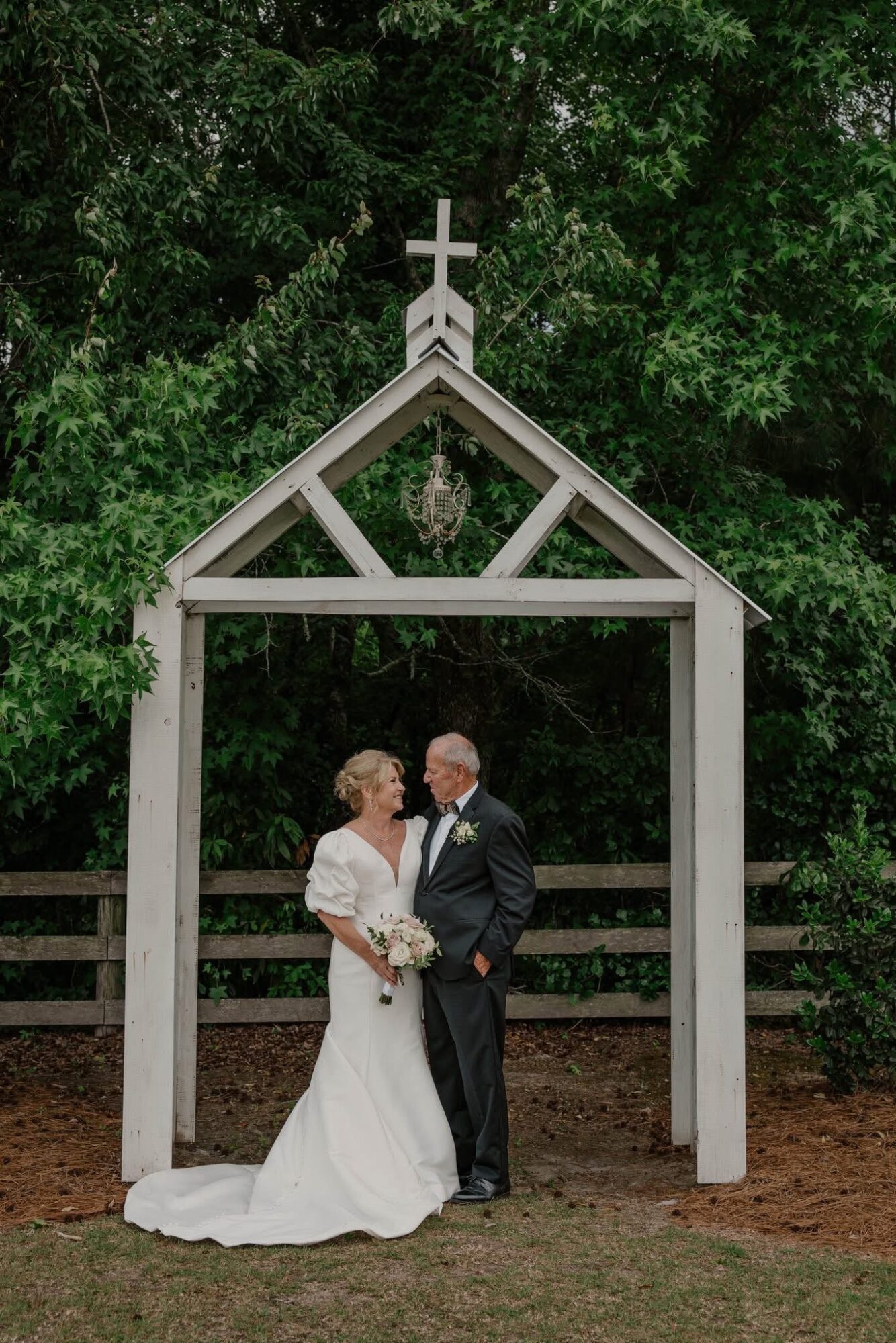 A bride and groom stand under a white wooden archway outdoors, surrounded by green trees, holding hands and smiling.