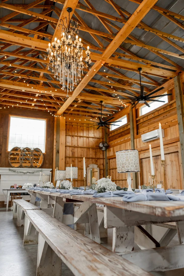 Interior of a rustic barn with a chandelier, wooden beams, and a long table set for dining.