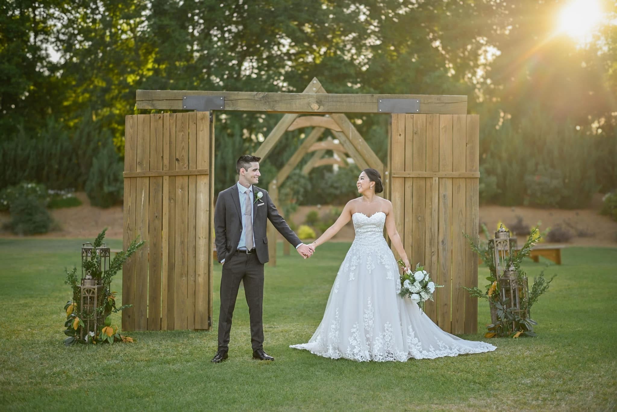 A bride and groom holding hands outdoors in front of a wooden arch, with sunlight and trees in the background.