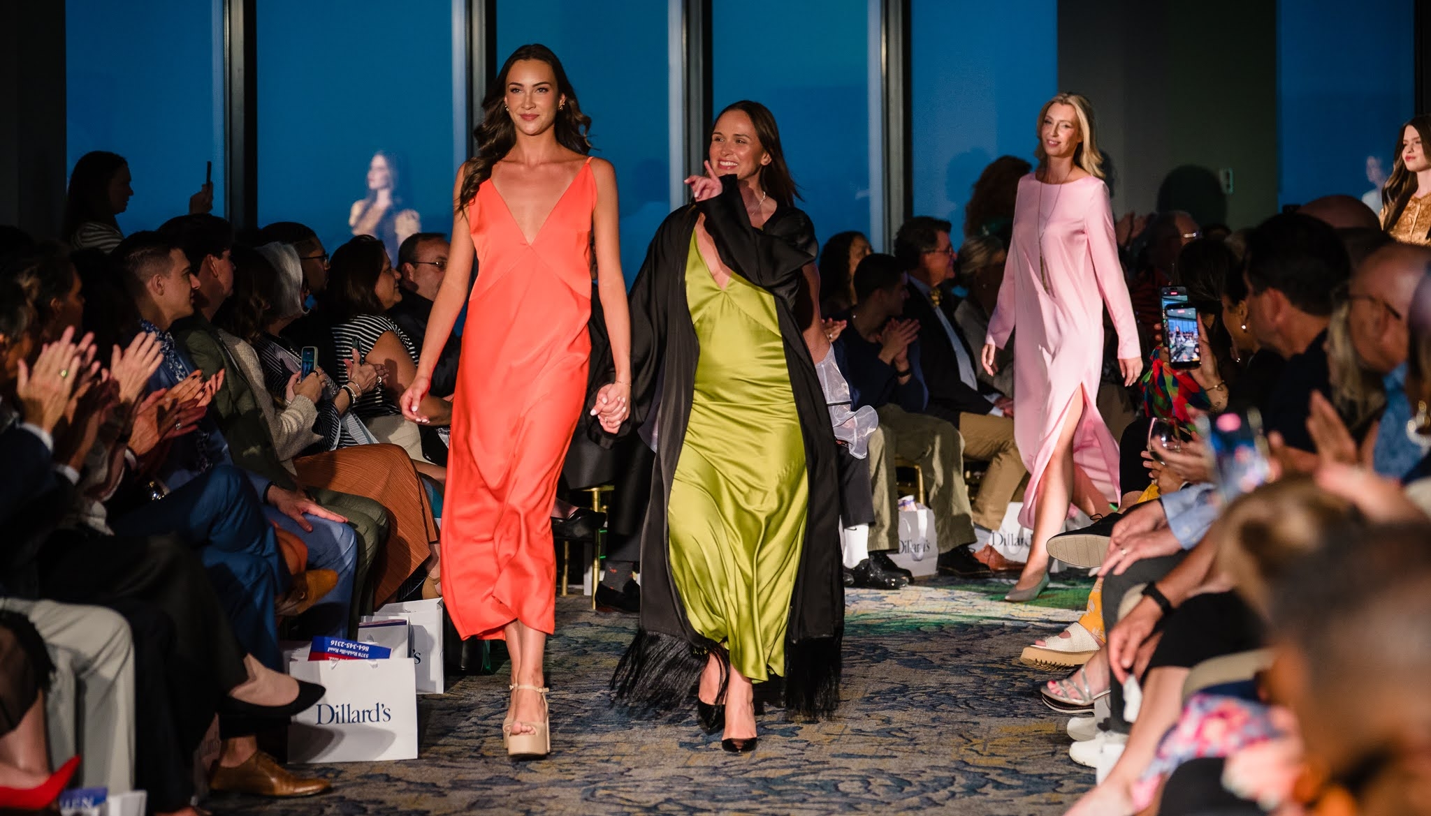 Models walk down runway in colorful dresses, audience seated on both sides clapping, fashion show setting.