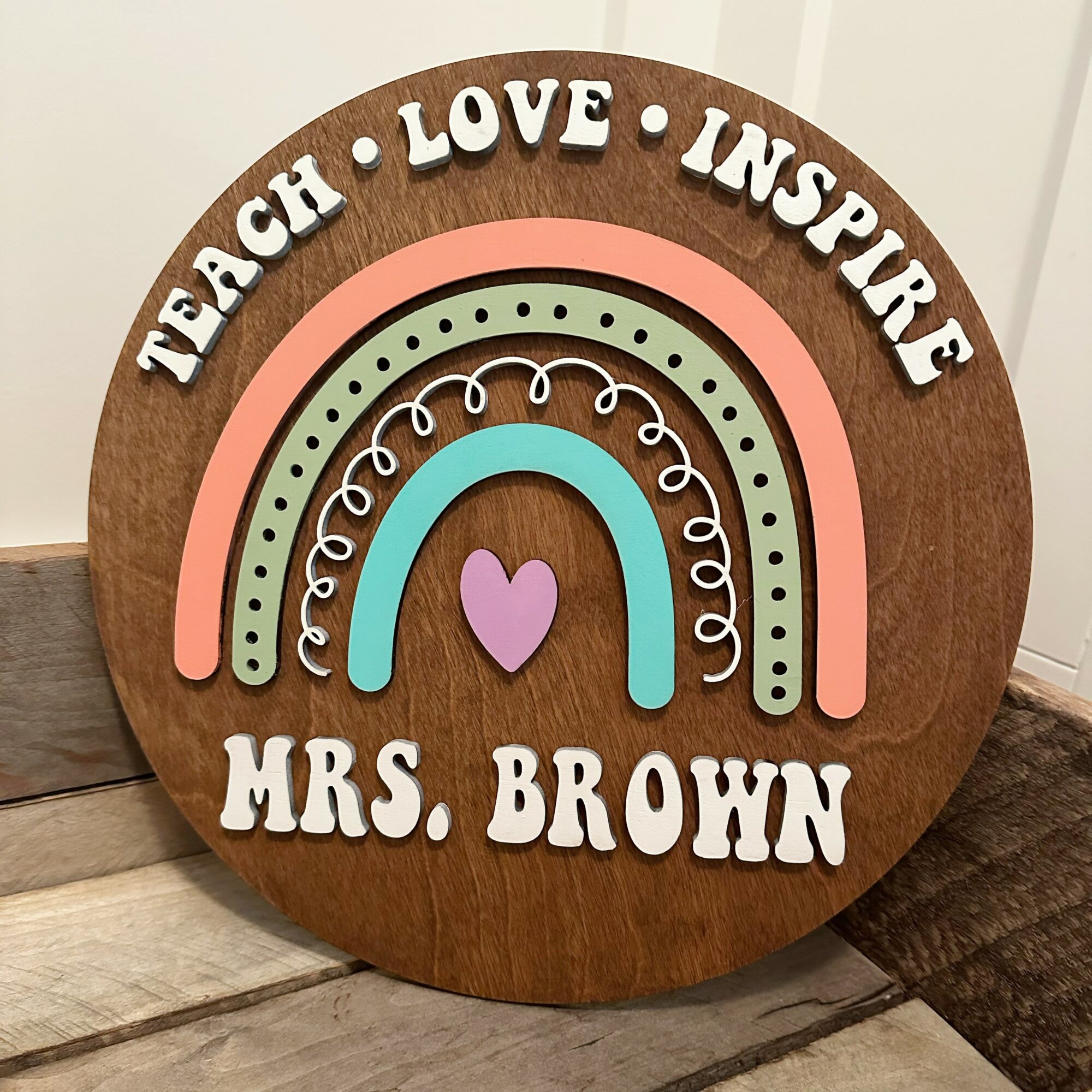 Decorative wooden sign with rainbow and heart, displaying words 'Teach Love Inspire' and 'Mrs. Brown'.