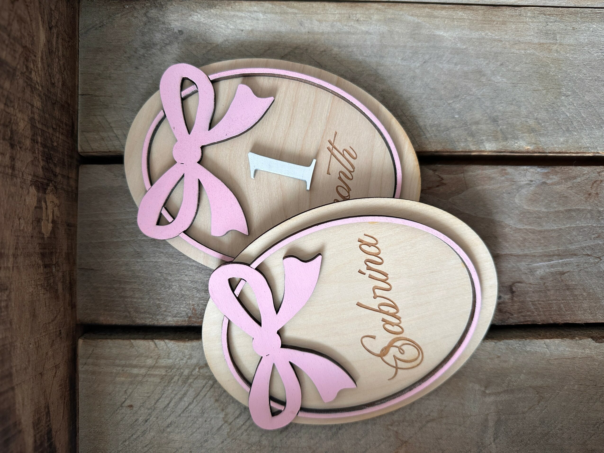 Two oval wooden signs with pink bows and the number 1 and the word 'April' written on them, placed on a wooden surface.