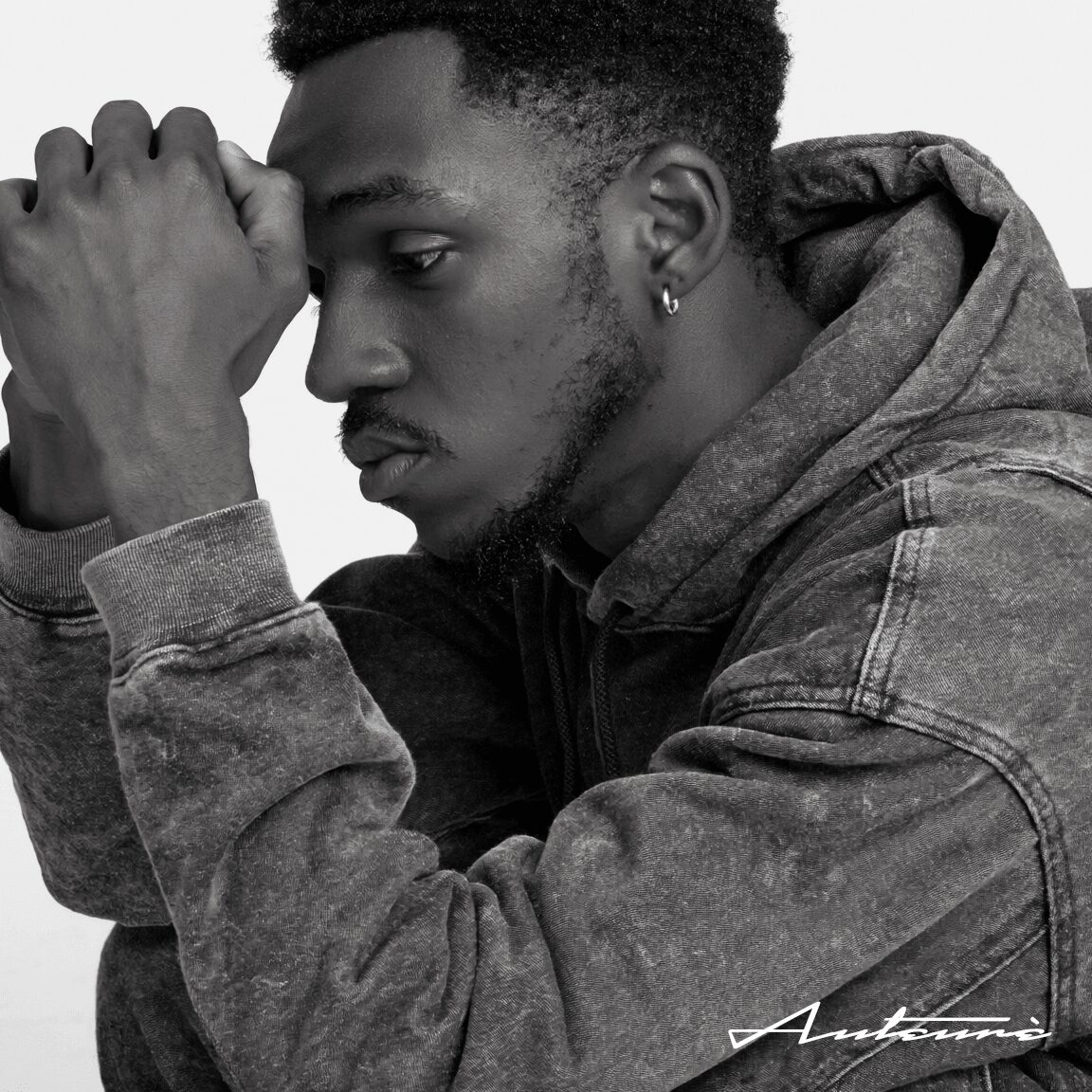 A young man with short curly hair and earrings, resting his head on his hand, wearing a hoodie and denim jacket.