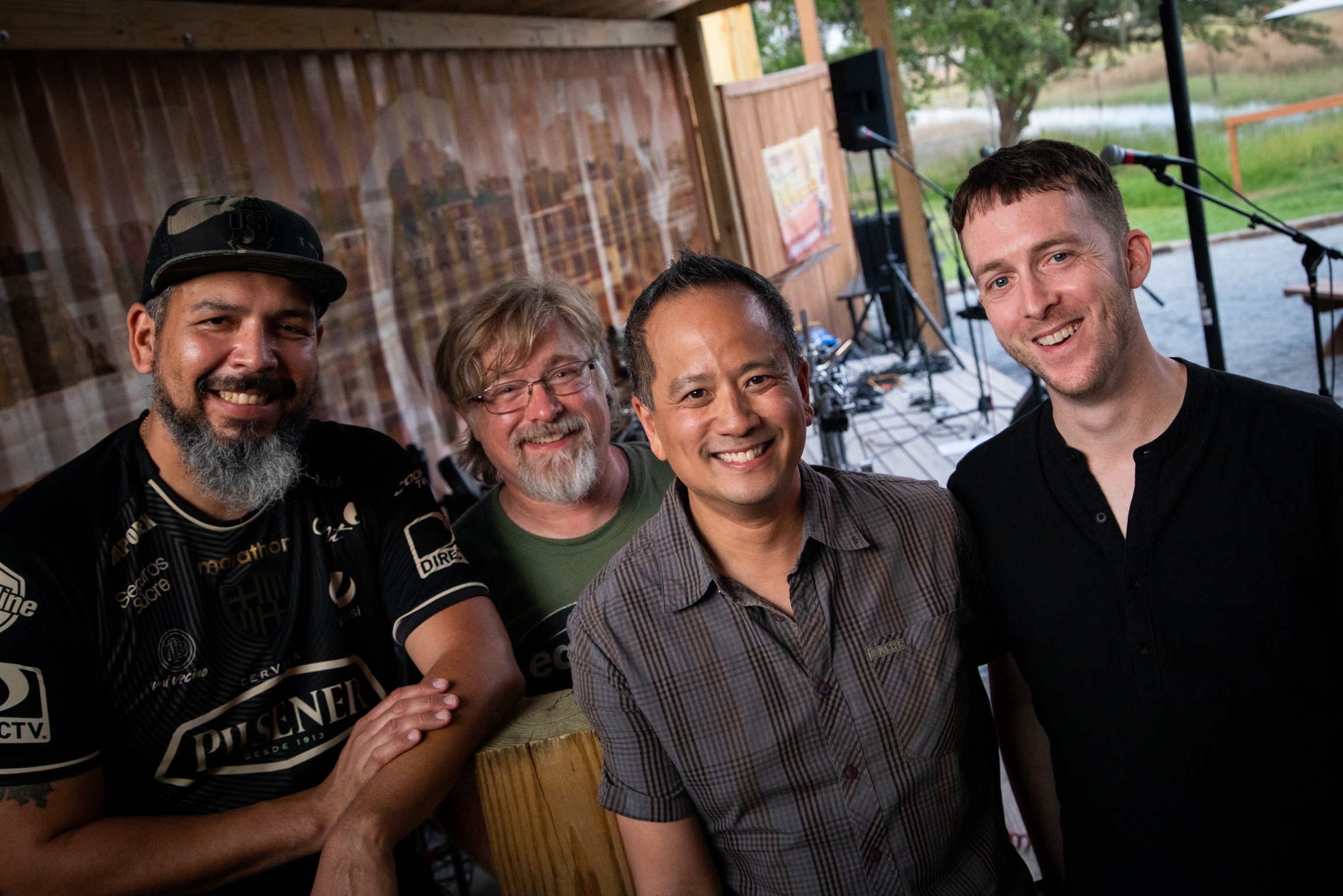 Four people smiling and standing together outdoors near a wooden structure, with a stage and musical equipment in the background.