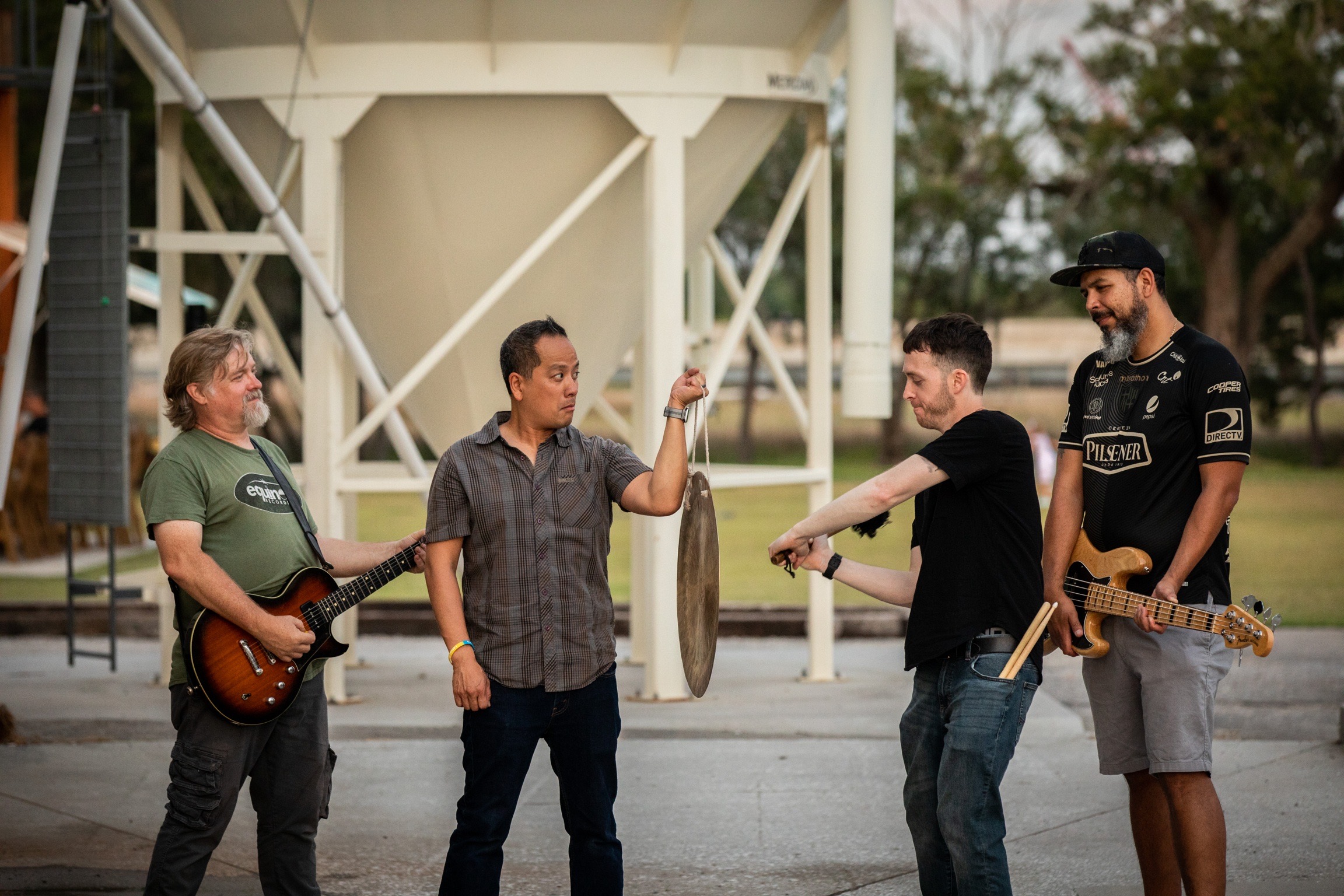 Four men with musical instruments standing outdoors near a large white structure, engaging in conversation and playing music.