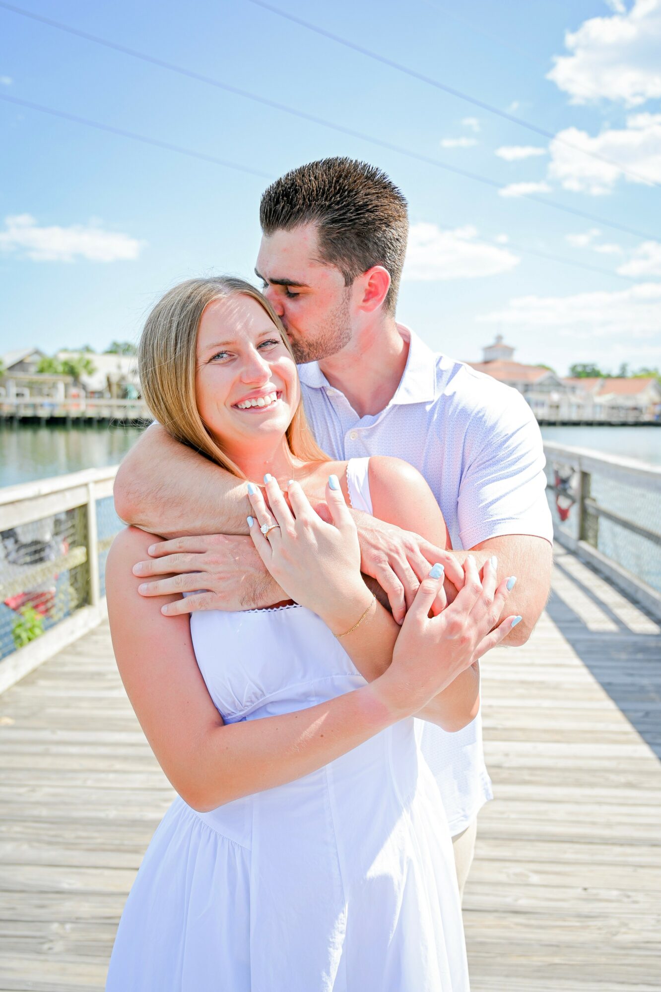 A couple embraces outdoors on a wooden dock, smiling, with a river and buildings in the background.