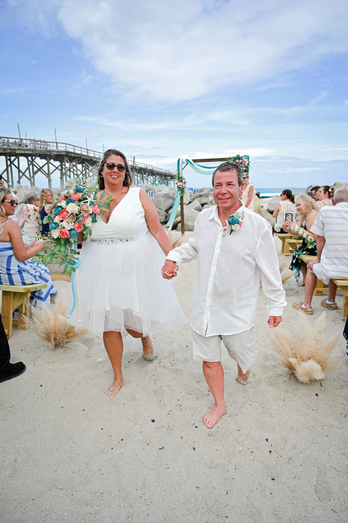 A couple walking barefoot on a beach, holding hands, with a wedding arch and guests in the background.