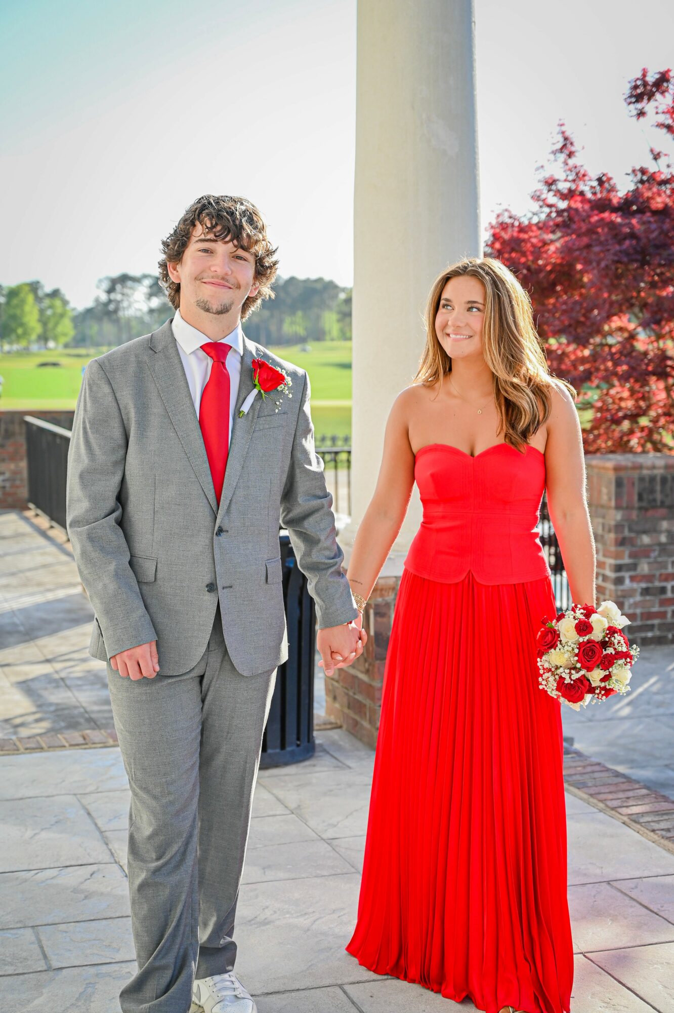 A couple holding hands outdoors, the man in a gray suit and red tie, the woman in a red dress holding a bouquet.