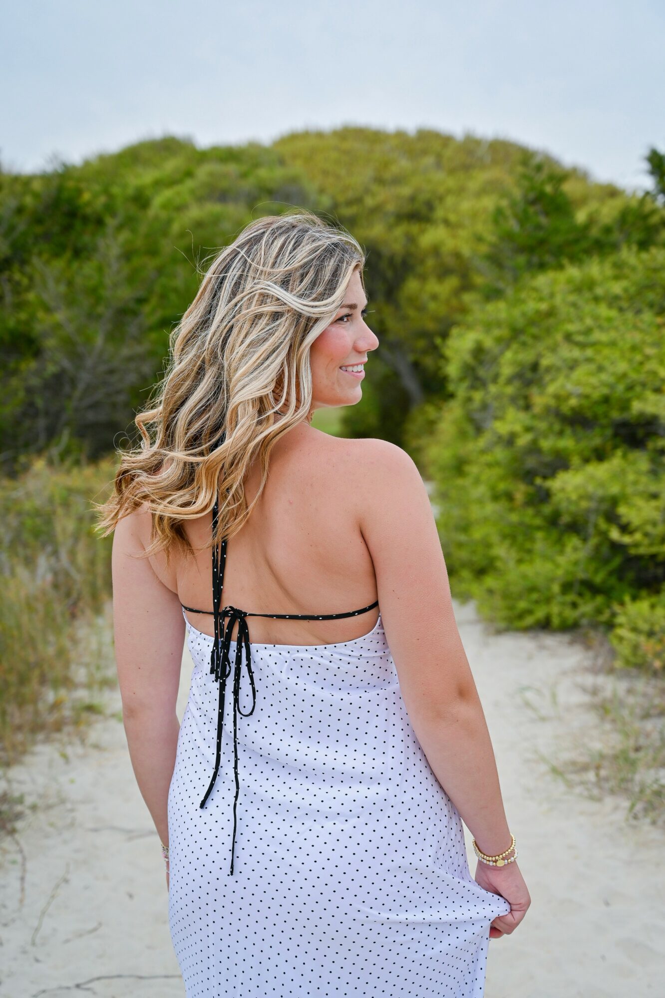 Woman with wavy blonde hair in a white dress with black straps standing outdoors on a sandy path, green trees in background.