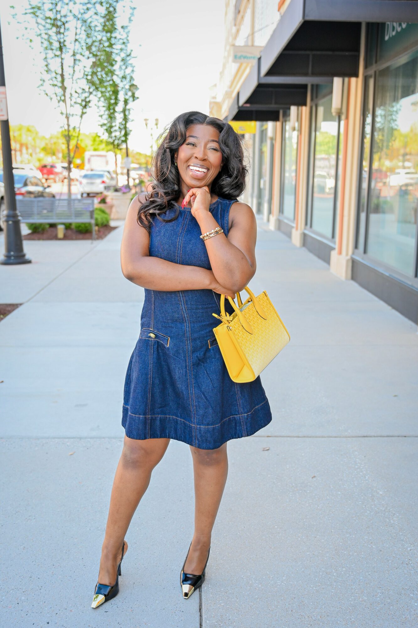 Young woman with curly hair smiling, standing on sidewalk with shopping bag, wearing denim dress and heels.