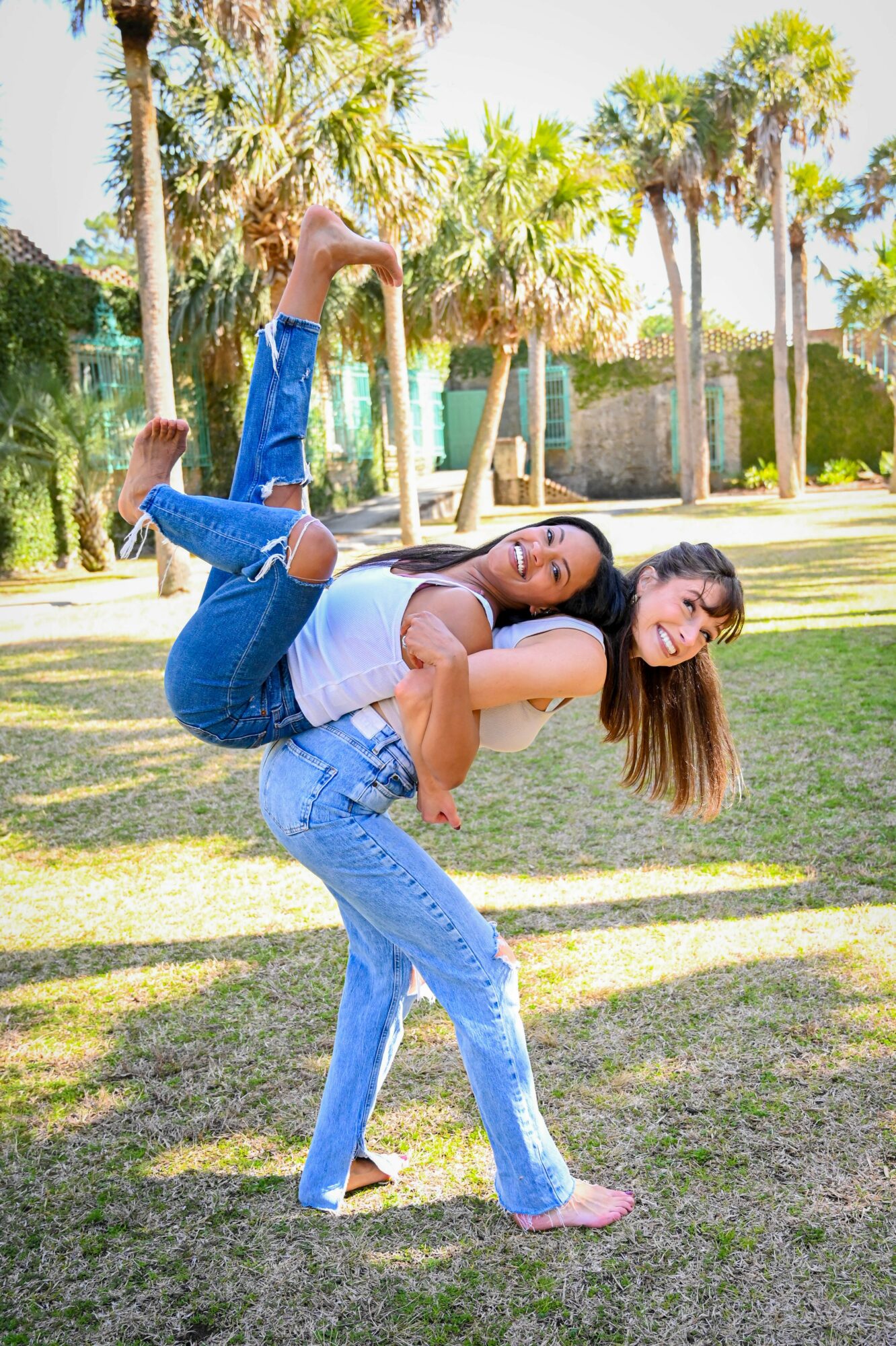 Two women outdoors, one carrying the other on her back, both smiling, with trees and a fence in the background.