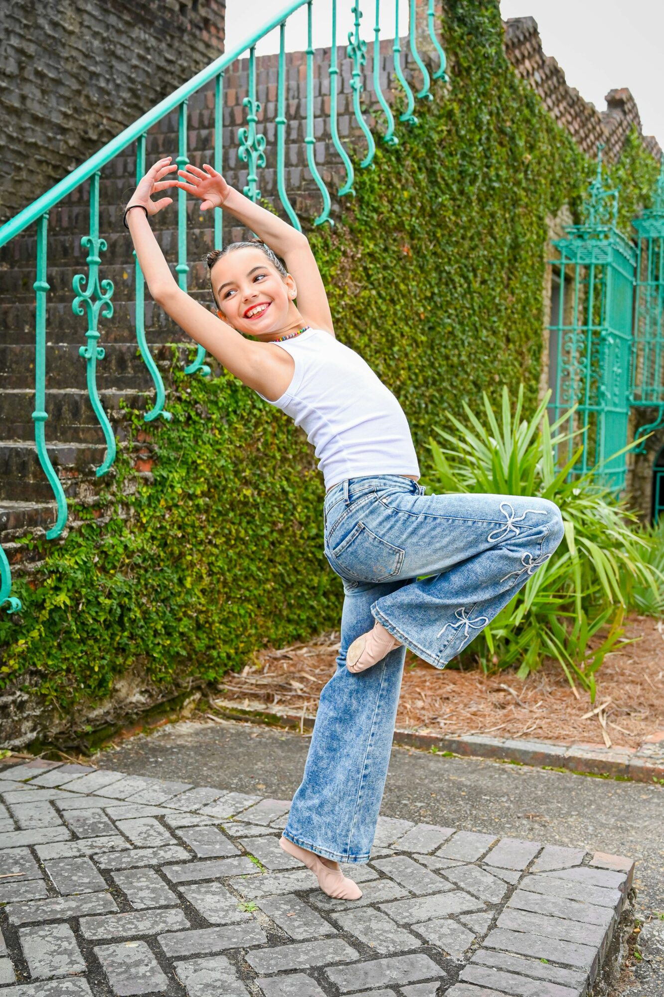 Young woman in white top and jeans poses outdoors near a green hedge and blue railing, smiling and stretching her arms.