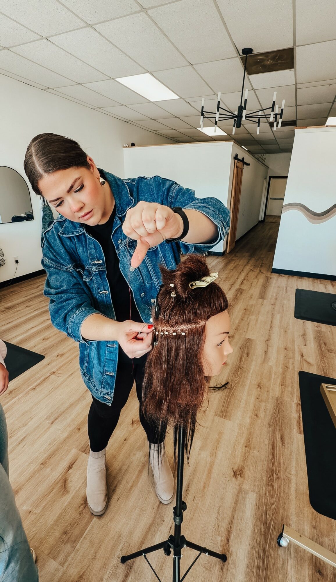 Woman practicing hairstyling on mannequin head in a salon, wooden floor, ceiling light, and modern decor.
