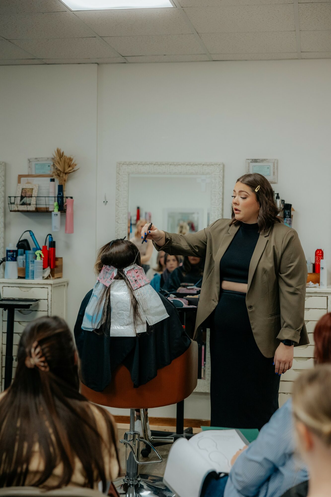 Woman teaching a group of children in a classroom, pointing at a mirror, with shelves and supplies in background.
