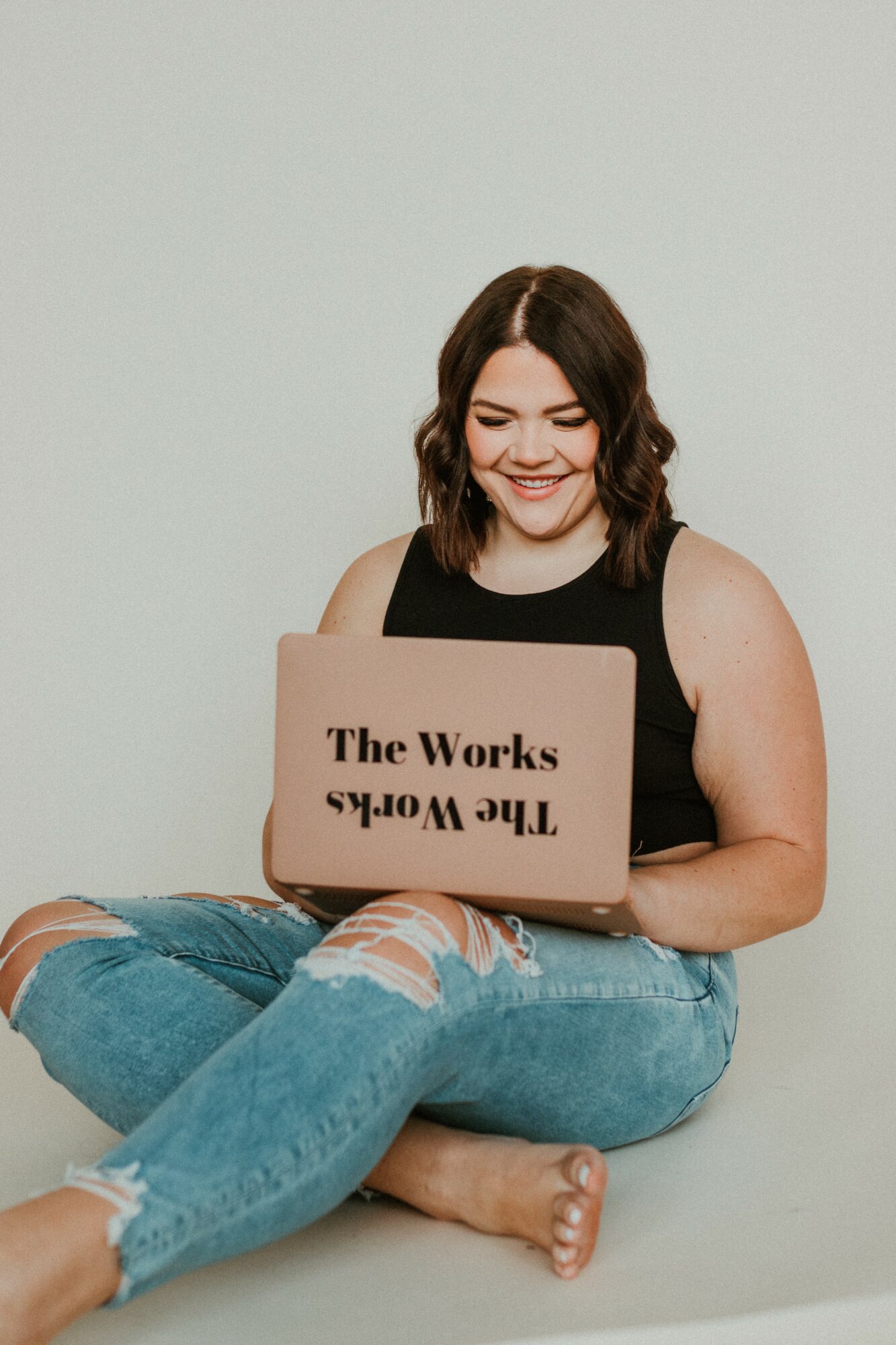 Young woman sitting cross-legged, smiling, holding a laptop with upside-down text, against a plain light background.