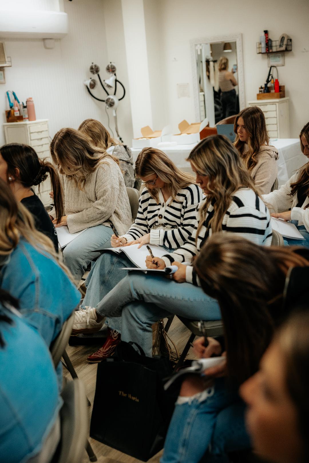 Group of women sitting in a room, some taking notes, with shelves and a mirror in the background.