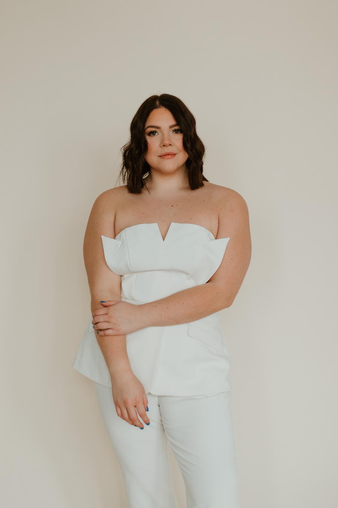 Woman with shoulder-length dark hair in a white strapless outfit, standing against a plain background.