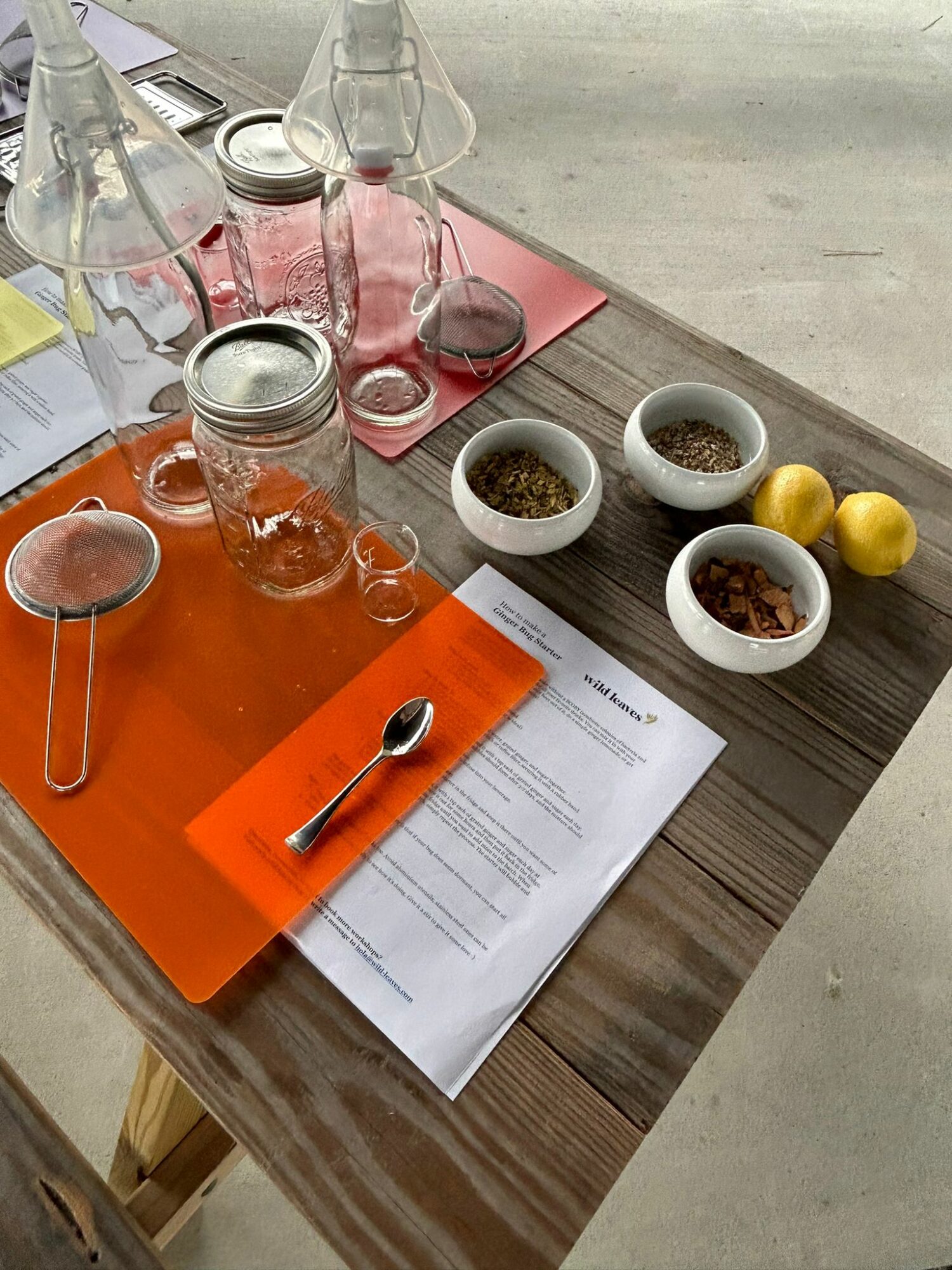 Table with glass bottles, jars, bowls of herbs, lemons, a spoon, and a paper with an orange cover.