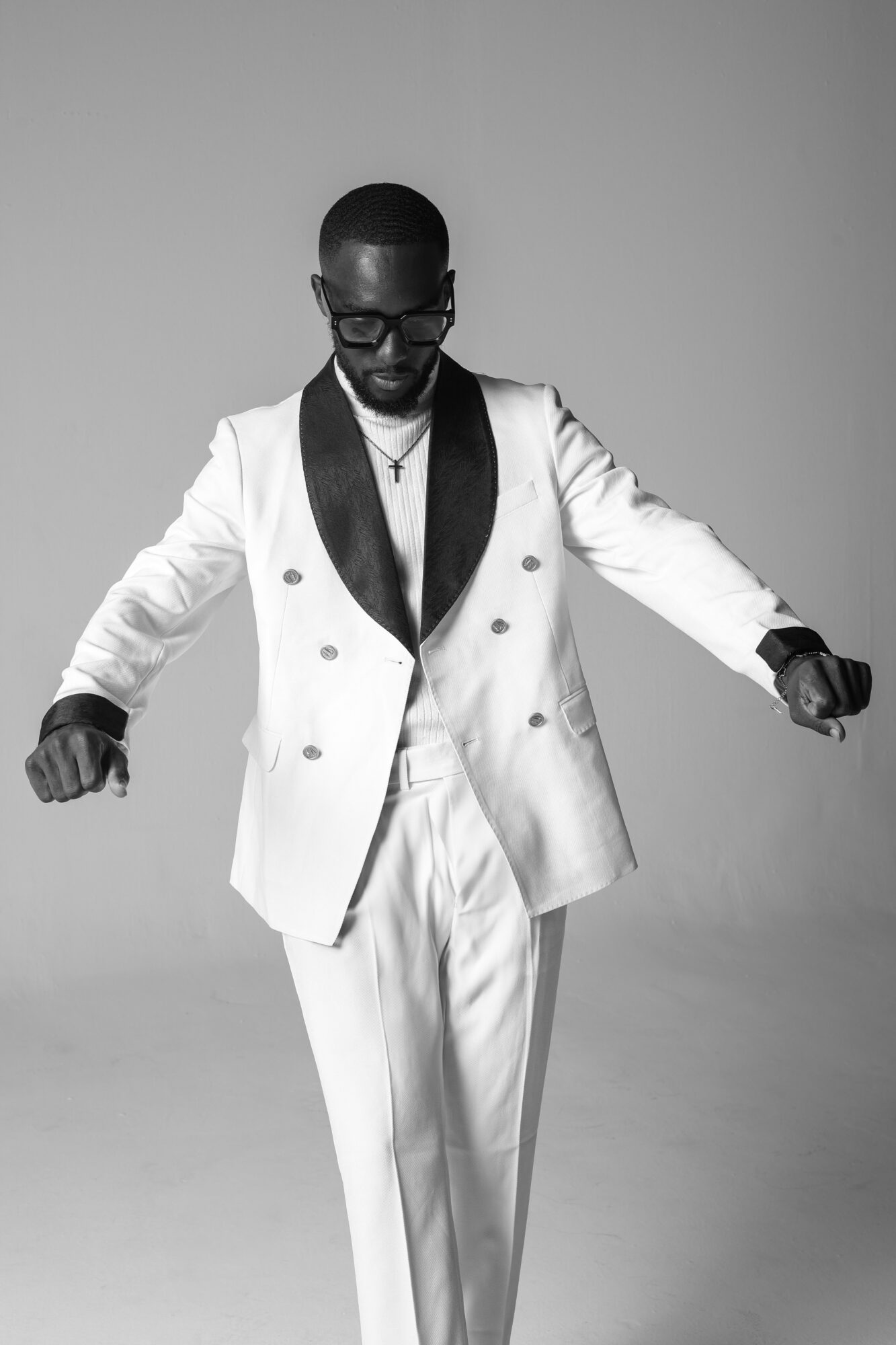 Man in a white suit with black lapel, glasses, and a cross necklace, standing with arms outstretched, looking down.