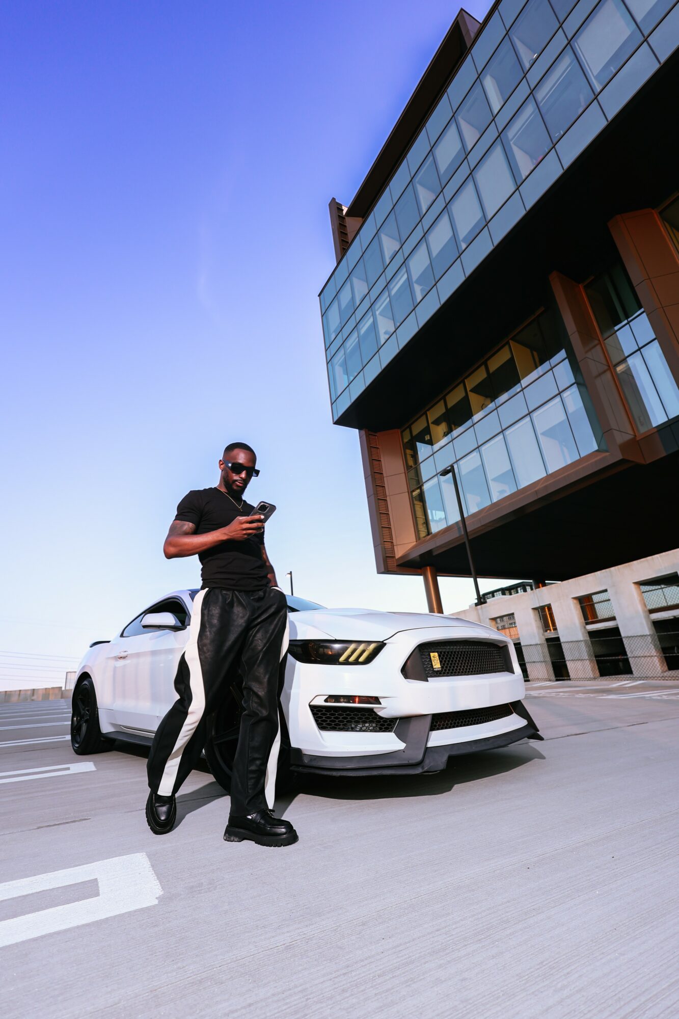 Man standing next to a white sports car, looking at his phone, in front of a modern building with glass windows.