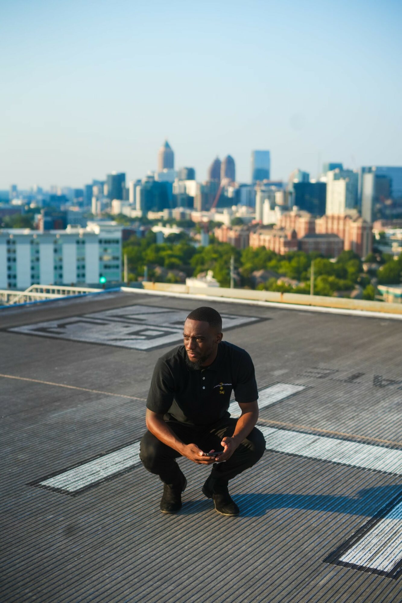 Person crouching on rooftop with city skyline in background, clear sky, daytime.