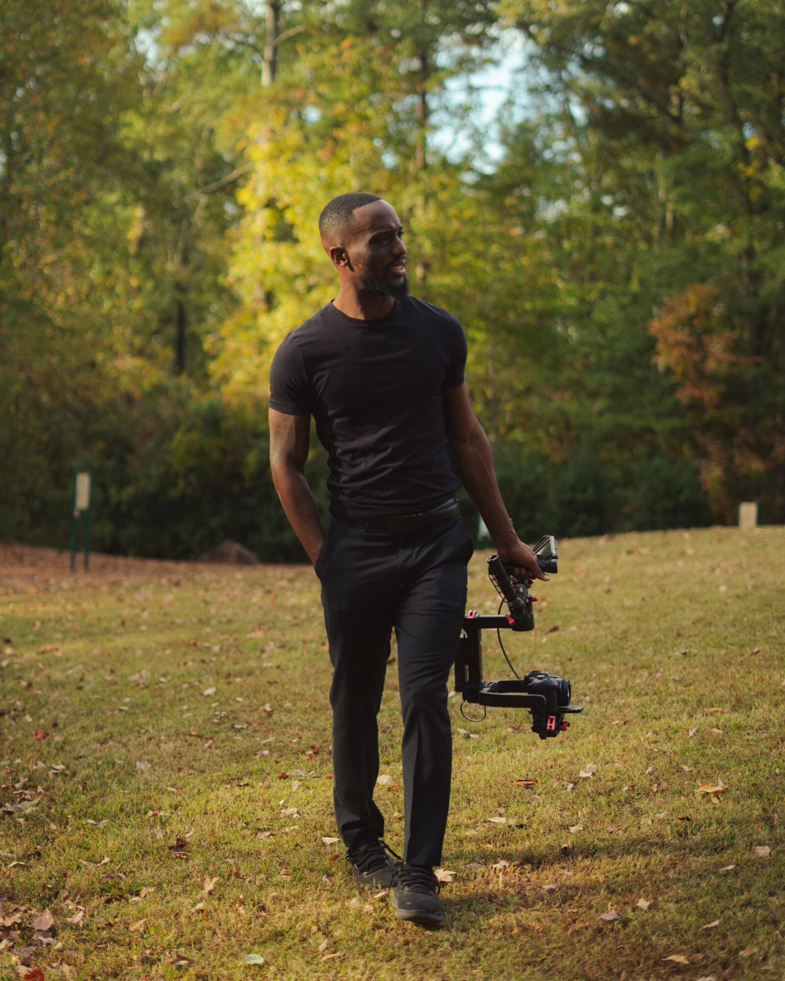 Man standing outdoors in a grassy area with trees, holding a camera stabilizer, wearing a black t-shirt and pants.