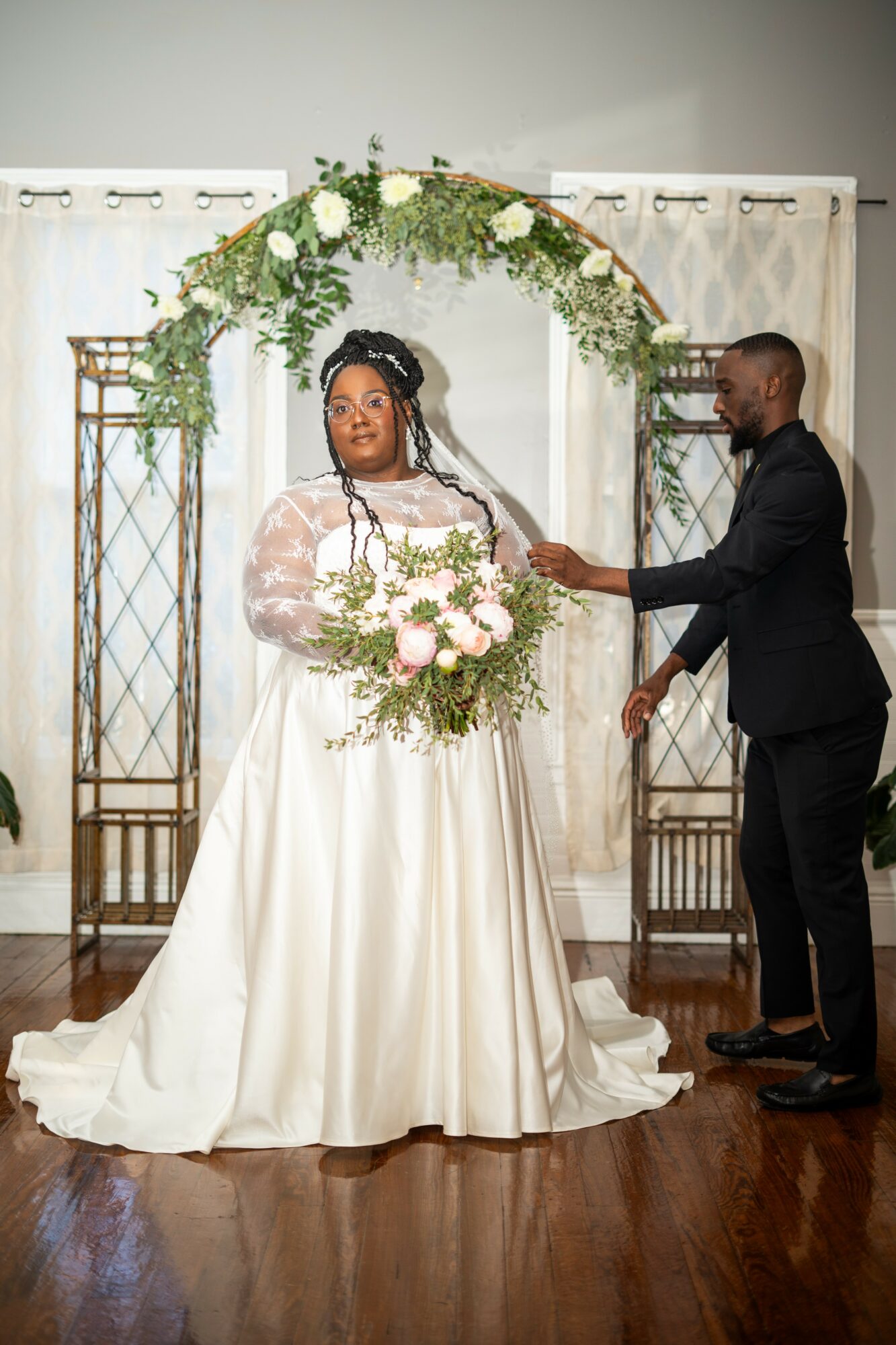Bride in a white gown holding a bouquet, groom in a black suit, standing under a floral arch, indoors.